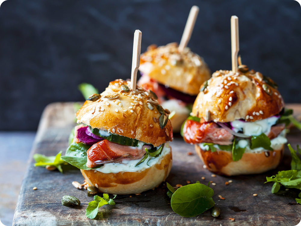 An aged wooden cheese board with mini artisan sliders complete with a large wooden toothpick. The sliders consist of sesame seed buns, smoked king salmon, spring greens, sliced cucumber, and fresh tarter sauce.