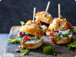 An aged wooden cheese board with mini artisan sliders complete with a large wooden toothpick. The sliders consist of sesame seed buns, smoked king salmon, spring greens, sliced cucumber, and fresh tarter sauce.